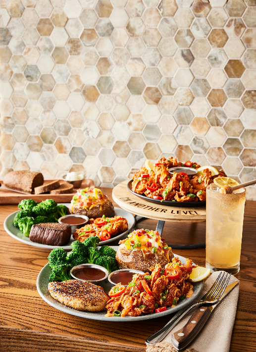 Outback chicken and steak entrees and appetizer served with cocktail on a wood counter with neutral tile backsplash. 